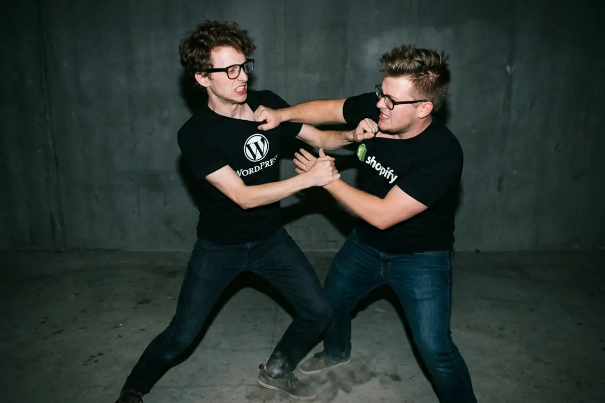 a couple of web designers fighting, one wearing a wordpress t-shirt and another one wearing a shopify t-shirt